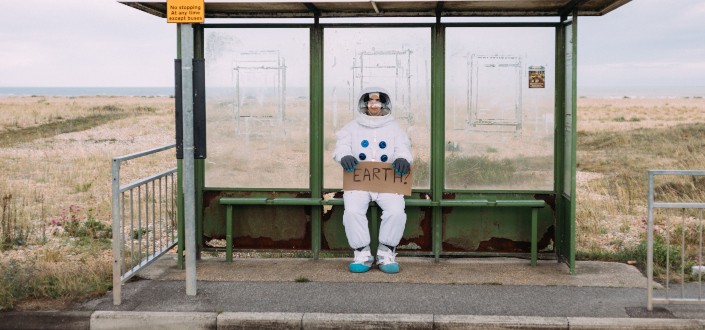 Astronaut-attired person holding a sign _Earth_ at a waiting shed.